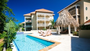 A hotel with a swimming pool, lounge chairs, and a thatched-roof structure. - Lawson Rock - Yellowfish 211 2 Bedroom Condo by RedAwning (Roatan)