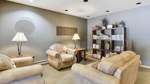 Uma sala de estar com sofá bege, mesinha de madeira, abajur, estante de livros e um objeto decorativo na parede.
