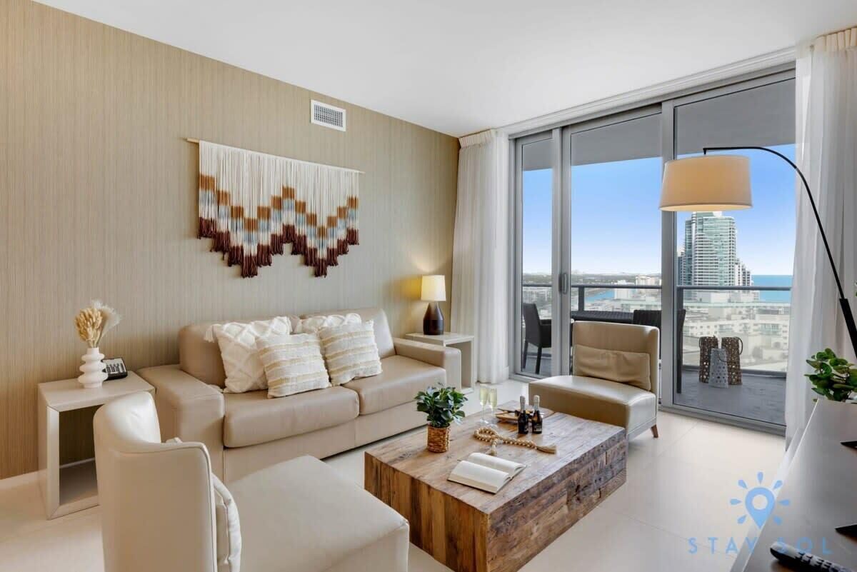 A modern hotel room with a large window offering a city view, a beige sofa, a wooden coffee table, and a decorative wall hanging.