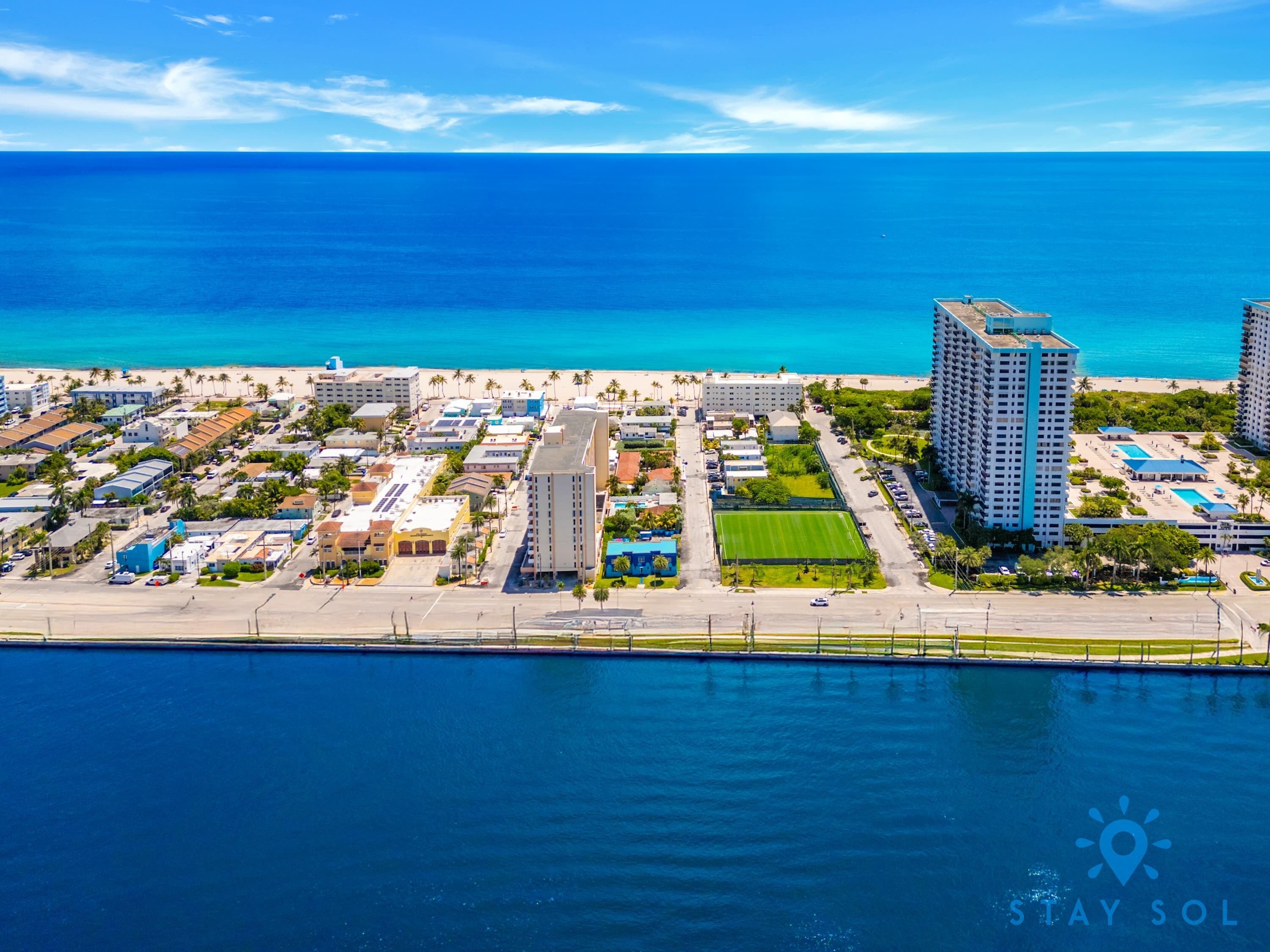An aerial view of a coastal resort with beachfront hotels, a marina, and a clear blue sky.