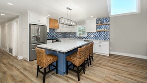 A modern kitchen with white cabinets, a large island with bar stools, a blue backsplash, and a wooden ceiling.