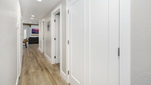 A hallway with white walls and wooden flooring, leading to a room with a gray sofa and a painting on the wall.