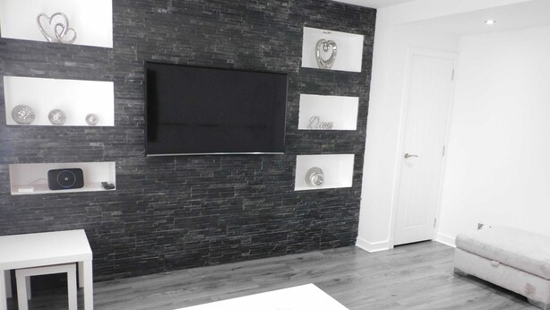 A modern living room with a flat-screen TV mounted on a brick-patterned wall, white furniture, and decorative items.