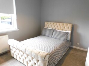 A bedroom with a tufted headboard, a bed with striped bedding, a radiator, and a window with a view of greenery. - Beautiful 3 bedroom home with large garden. 35 mins to Glasgow & Edinburgh. (Shotts)