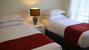 Two beds with white bedding and red accents, a bedside lamp, and a window with sheer curtains. - Wagga Showground Villas (Turvey Park)