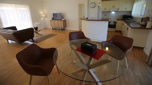 A modern living room with a glass coffee table, brown leather chairs, a sofa, and a TV. The kitchen is visible with wooden cabinets and a clock. - Wagga Showground Villas (Turvey Park)
