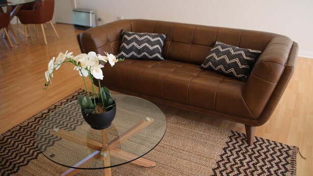 A brown leather sofa with two patterned pillows, a glass coffee table with a potted orchid, and a herringbone-patterned rug. - Wagga Showground Villas (Turvey Park)