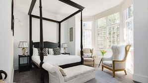 Un lit à baldaquin avec un cadre sombre, une petite table de chevet avec une lampe, une chaise et une vue sur l’extérieur grâce à de grandes fenêtres.
