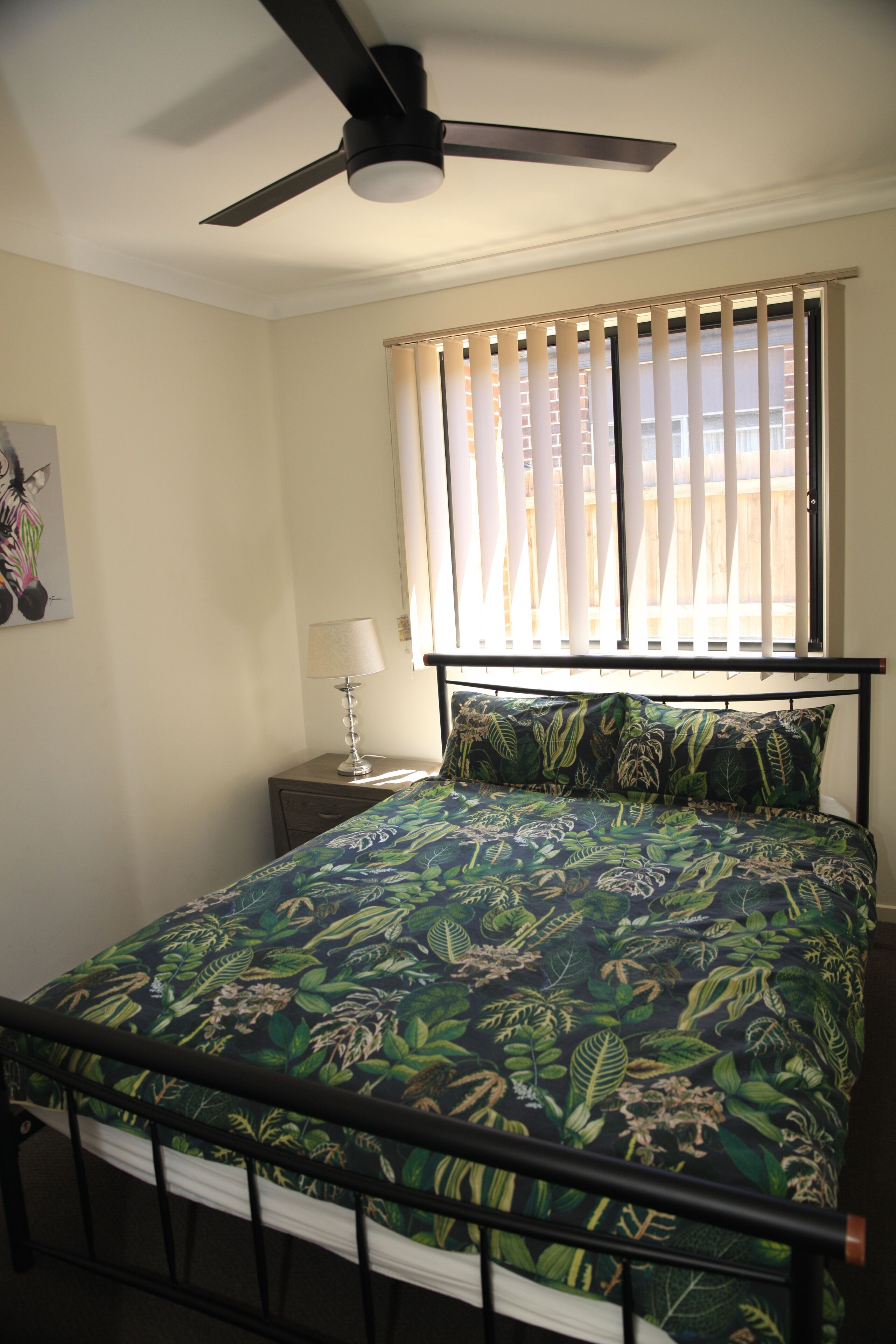 A bedroom with a bed, a ceiling fan, a window with blinds, a bedside table with a lamp, and a wall-mounted artwork.