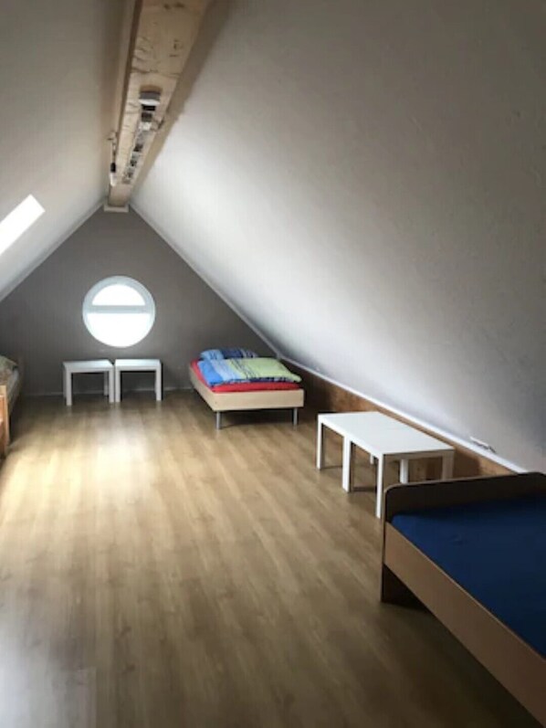 Une petite chambre mansardée de forme triangulaire, avec un plancher en bois, une lucarne ronde, un lit, un bureau et un banc.