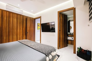 A modern bedroom with a large bed, wooden wardrobe, a flat-screen TV, and a bathroom visible through an open door. - NEW 1 BR w/pool and gym @ Desire (Puerto Vallarta)