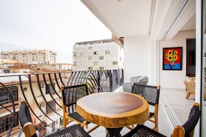A balcony with a round wooden table and black wicker chairs, overlooking a cityscape with modern buildings.