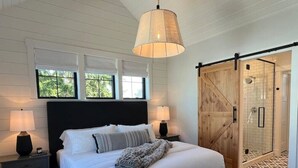 A bedroom with a large bed, two bedside tables, a bench, a hanging lamp, and a sliding door leading to a shower area. - New luxury home steps to downtown w/yr round heated pool (New Buffalo)