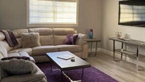 Ein Wohnzimmer mit einem beigen Sofa, einem Couchtisch, einem violetten Teppich, einem kleinen Beistelltisch und einem an der Wand montierten Fernseher.
