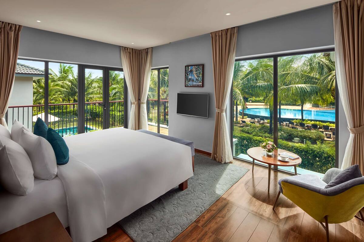 A modern hotel room with a large bed, a small round table, a yellow armchair, and a view of a swimming pool and palm trees.