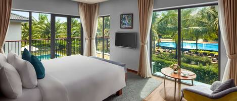 A modern hotel room with a large bed, a small round table, a yellow armchair, and a view of a swimming pool and palm trees.
