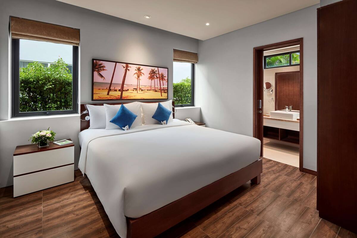 A modern bedroom with a large bed, a nightstand with a lamp, a painting of a beach scene, and a view of palm trees through the window.