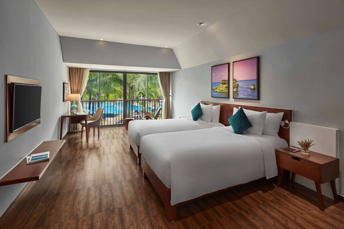 A hotel room with two beds, a TV, a desk with a chair, and a view of a pool and palm trees.