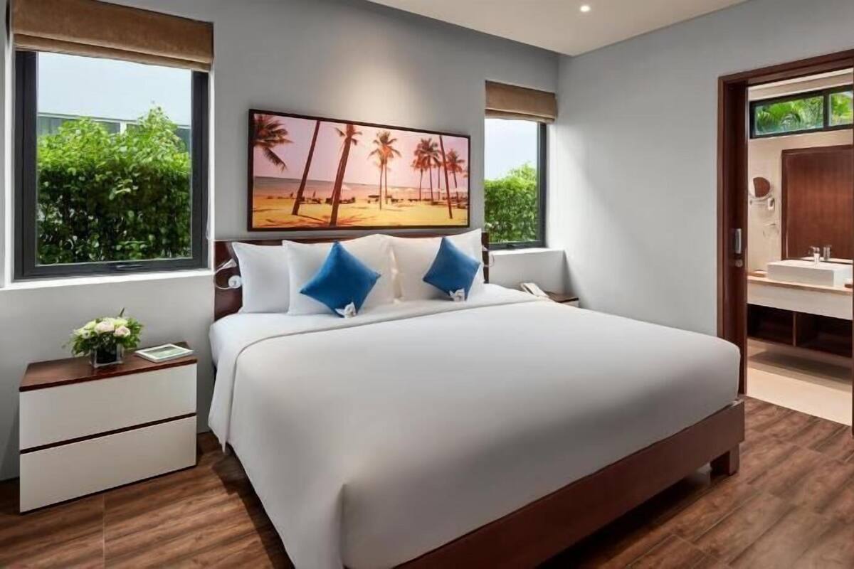 A bedroom with a large bed, bedside tables, a nightstand, a painting of a beach scene, and a bathroom visible through an open door.