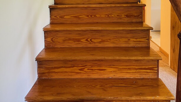 A wooden staircase with a handrail, leading to a landing with a door and a window with curtains.