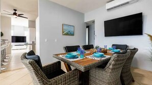 A dining area with wicker chairs, a wooden table set with blue plates and glasses, a flat-screen TV mounted on the wall, and a ceiling fan. - Parguera Paradise, Luxury Modern Beach House! (Lajas)
