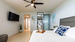 A bedroom with a bed, a ceiling fan, a flat-screen TV, and a door leading to a bathroom. - Parguera Paradise, Luxury Modern Beach House! (Lajas)