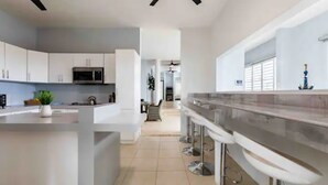 A modern kitchen with white cabinets, a central island with bar stools, and a microwave. - Parguera Paradise, Luxury Modern Beach House! (Lajas)