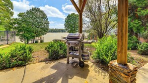 Ein Grill mit Holzbalkonüberdachung und Steinsockel, platziert in einem Garten mit Seeblick und Bäumen.