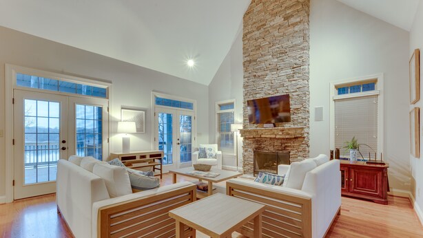 Ein geräumiges Wohnzimmer mit Kamin aus Stein, weißen Sofas, Holztischen und großen Fenstern mit Jalousien.