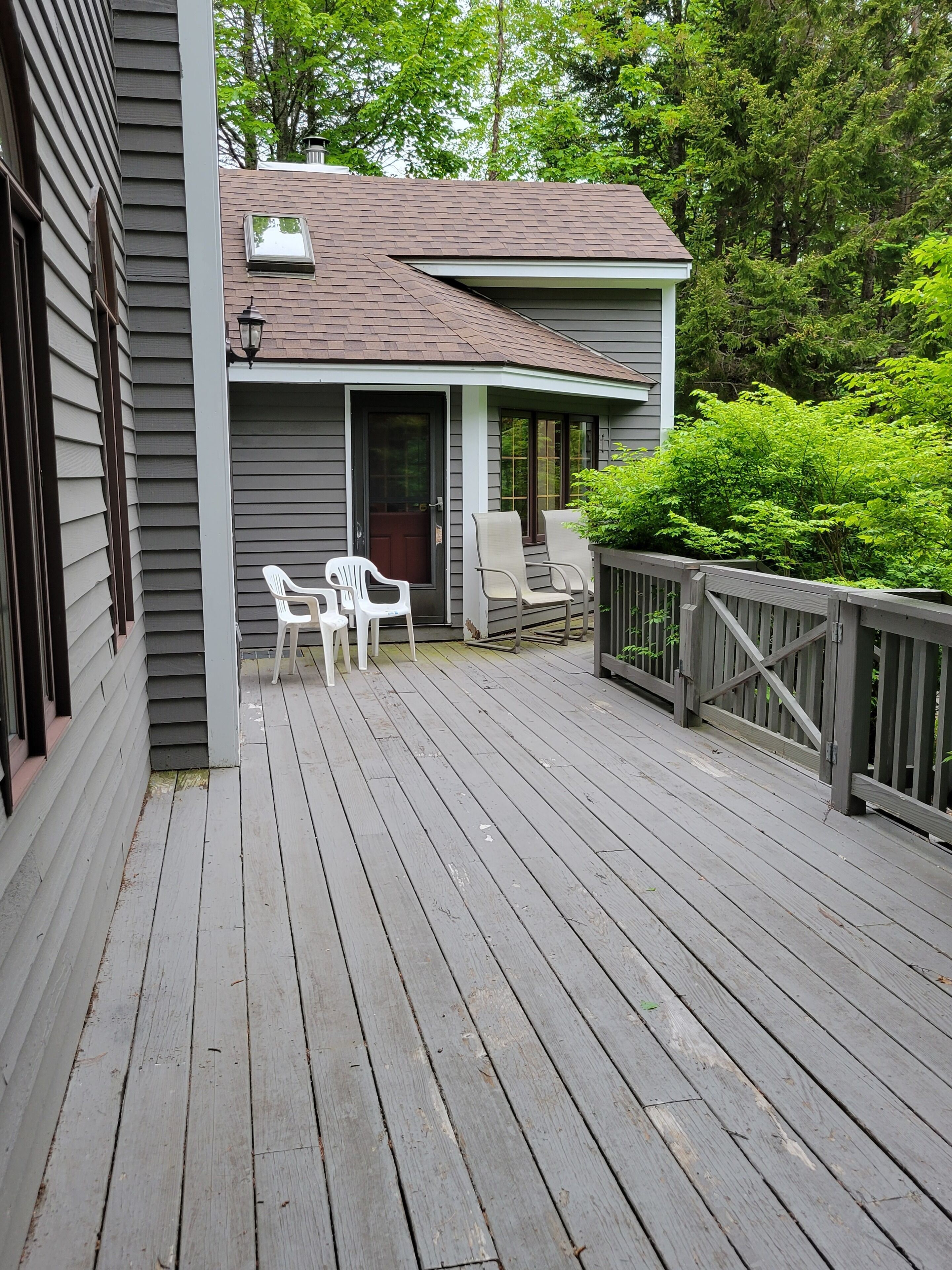 Eine Holzterrasse mit zwei weißen Plastikstühlen und einem kleinen Tisch, einem Holzzaun und einem Haus mit dunklem Dach und heller Außenverkleidung.