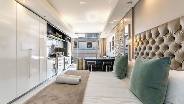 A modern hotel room with a large bed, a tufted headboard, a small kitchenette, and a view of the city. - Sleek Central Apartment | Wifi | Pool | Gym (Cape Town)