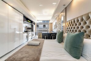 Une chambre d’hôtel moderne dotée d’un grand lit, d’une tête de lit capitonnée, d’une petite kitchenette et offrant une vue sur la ville.