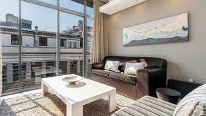 A modern living room with a large window, a sofa, a coffee table, and a painting on the wall. - Sleek Central Apartment | Wifi | Pool | Gym (Cape Town)