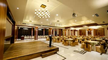 A spacious banquet hall with multiple round tables and chairs, a podium, and a wooden stage.