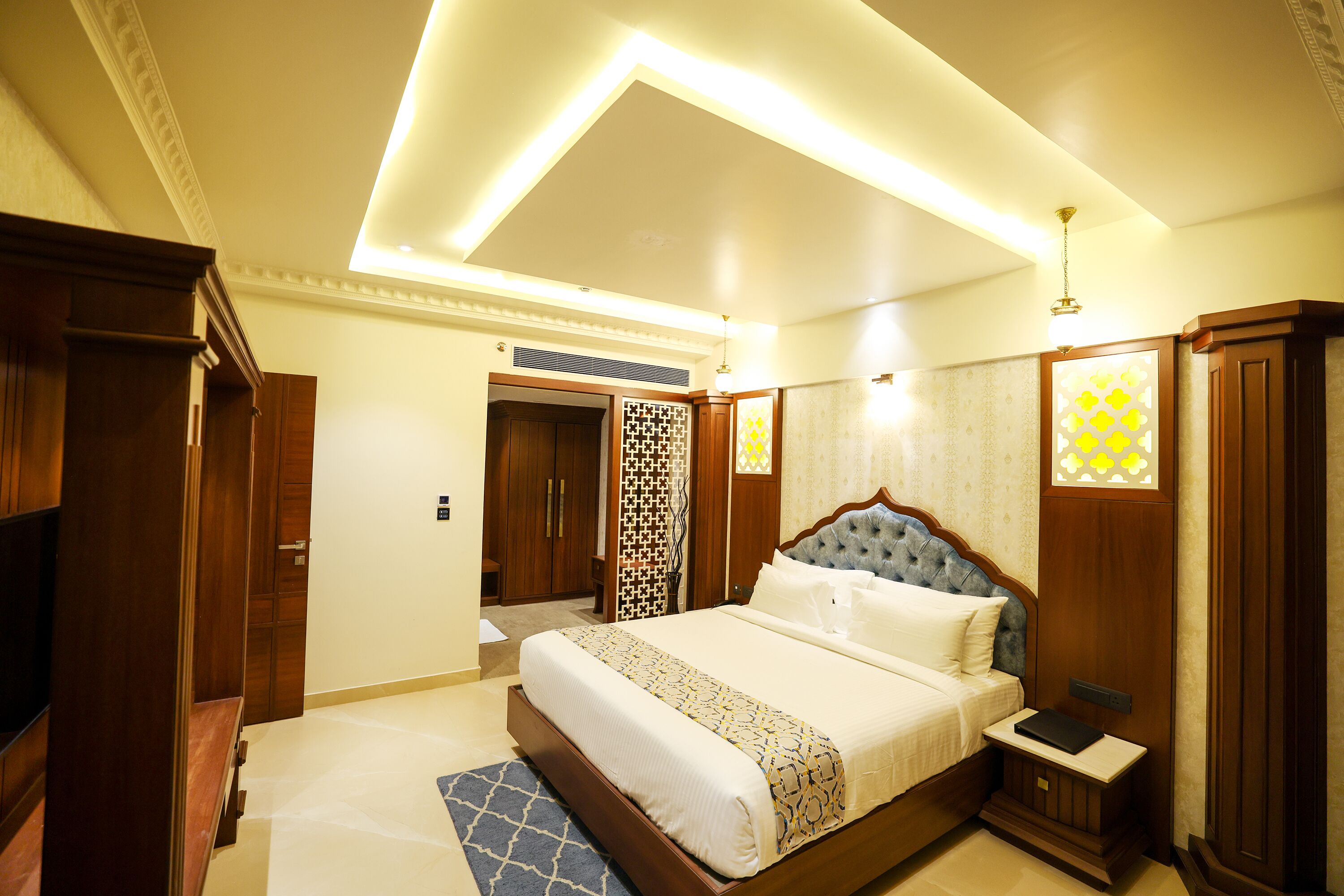 A well-lit bedroom with a large bed, wooden wardrobe, and a small bedside table.