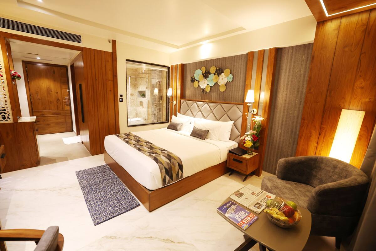 A hotel room with a large bed, a wooden headboard, a nightstand with a lamp, a seating area with a chair, and a table with a fruit bowl.