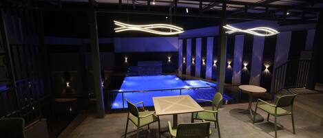 Un área de piscina interior con piscina azul, sillas verdes y una mesa.