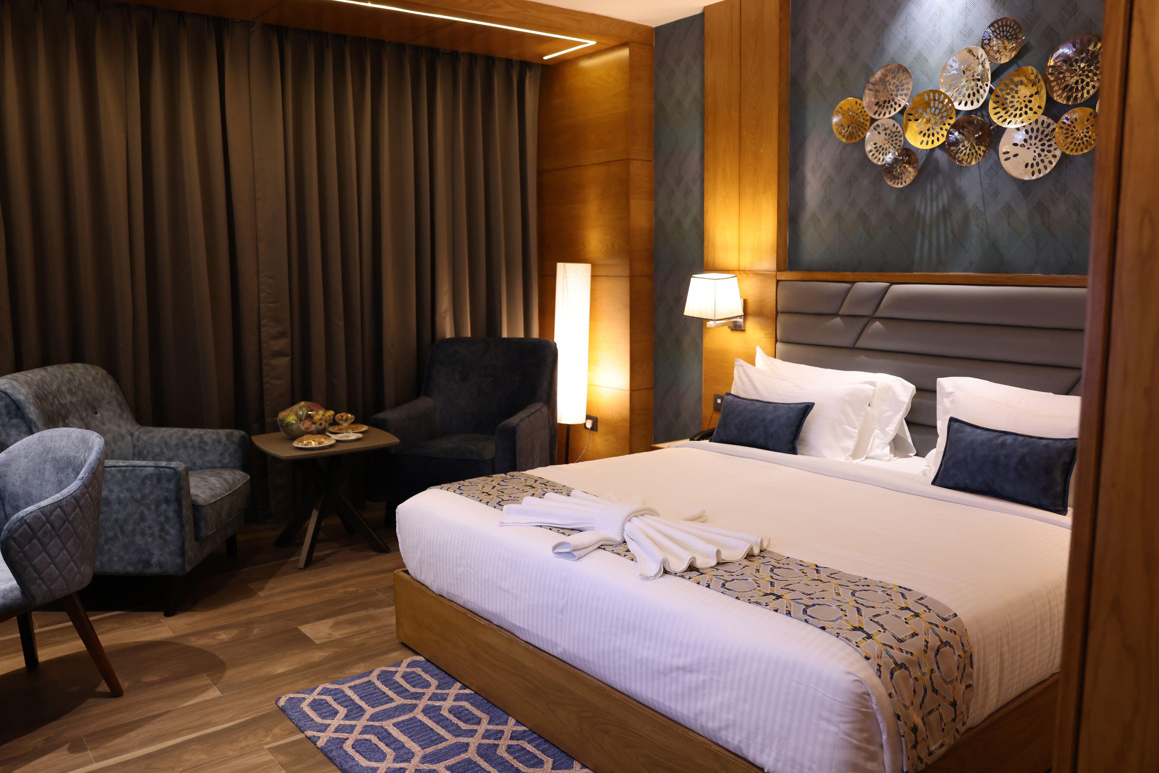 Quarto de hotel com cama, duas poltronas, uma mesinha com um prato de comida, uma luminária na parede e um objeto decorativo na parede.