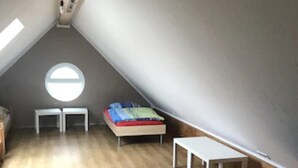 Une petite chambre mansardée de forme triangulaire, avec un plancher en bois, une lucarne ronde, un lit, un bureau et un banc.