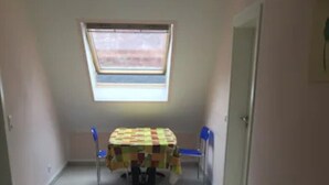 Une petite pièce avec un sol carrelé, une table recouverte d’une nappe à carreaux, deux chaises bleues et une fenêtre avec des stores.