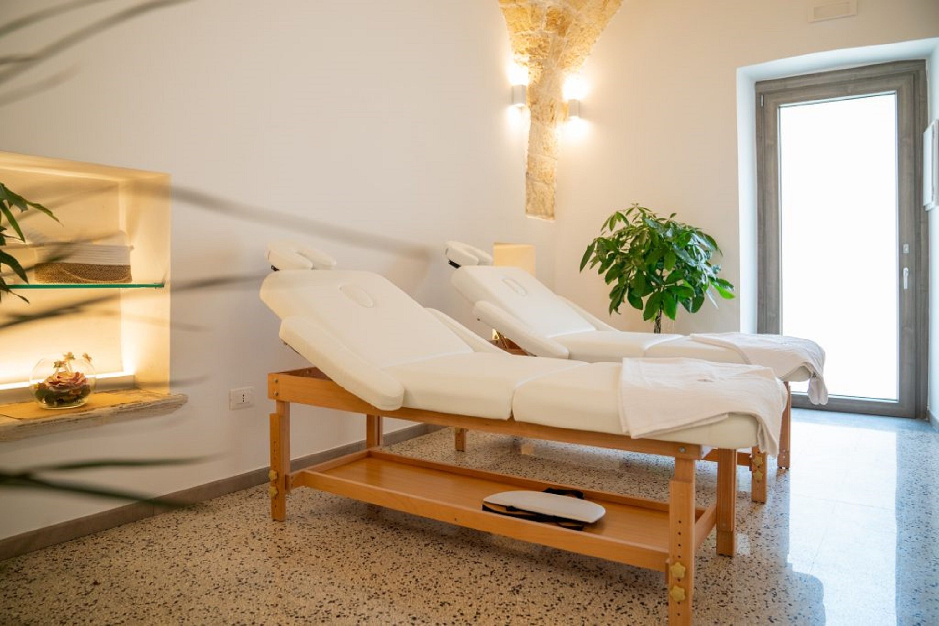 Ein Wellnessraum mit zwei Massagetischen, einer Steinwand, einer Pflanze und einer Tür.