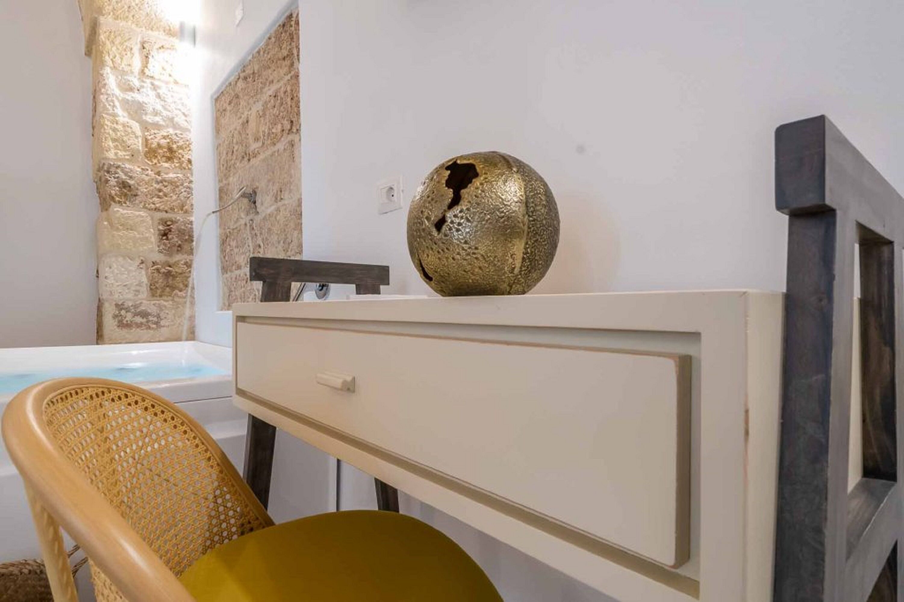 Une salle de bain avec un mur en pierre, une baignoire, un meuble-lavabo blanc agrémenté d'un bol décoratif doré et une chaise en osier.