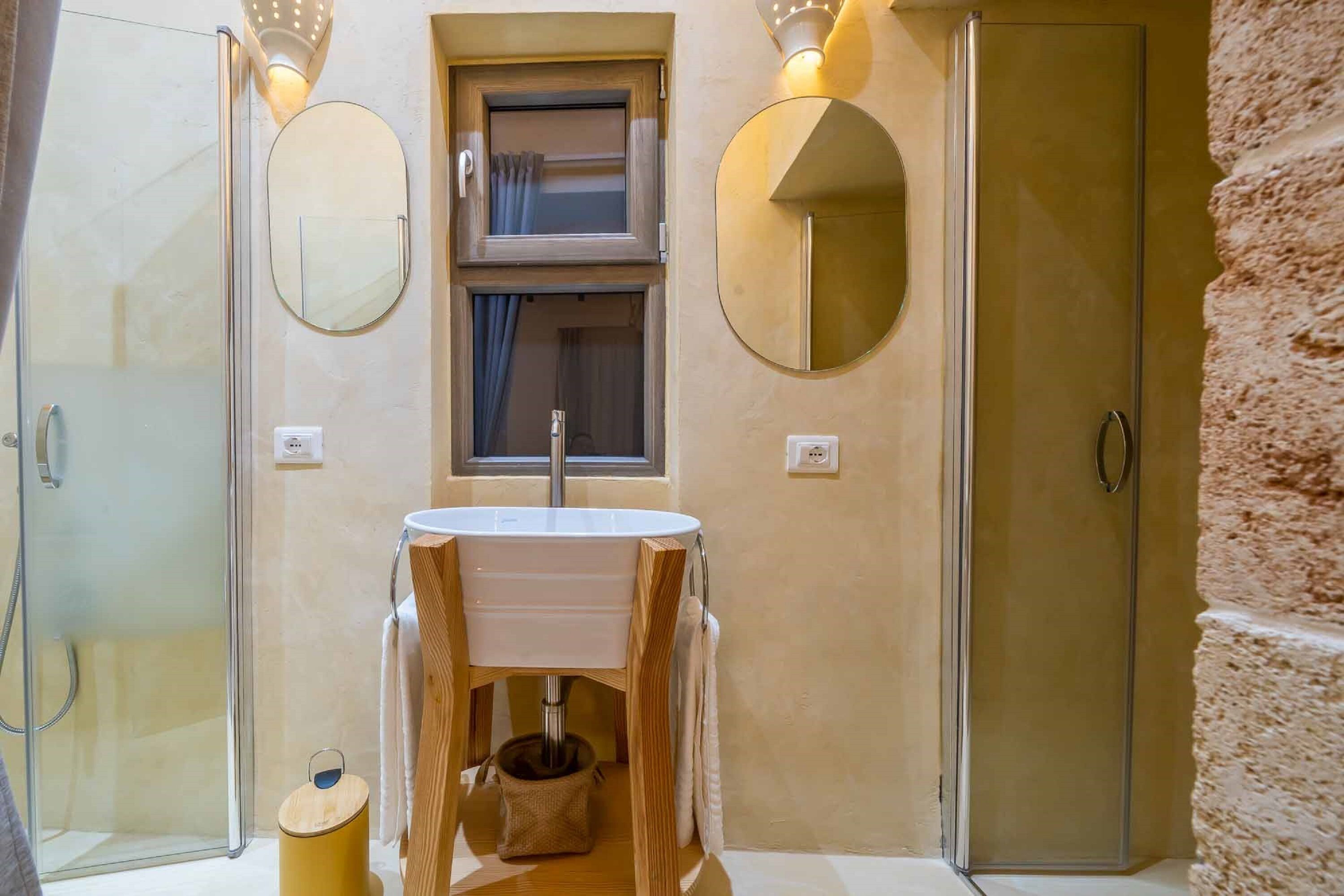 A bathroom with a wooden vanity, two wall-mounted mirrors, a window with curtains, and a shower area.