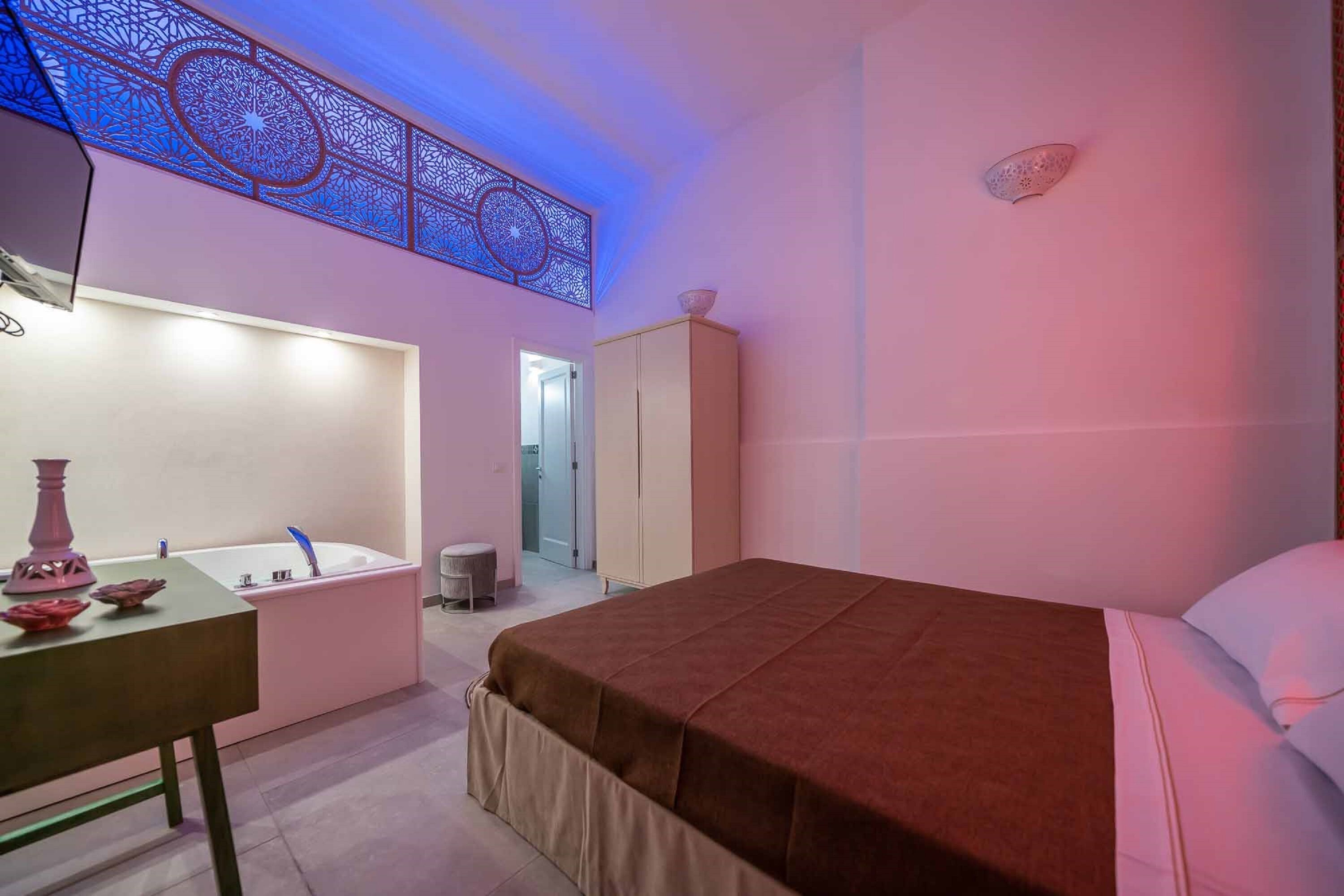 A bedroom with a bed, a nightstand, a vanity with a mirror, a bathtub, and a wardrobe.