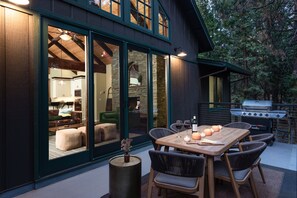An outdoor dining area with a wooden table and chairs, a grill, and a cozy interior visible through large glass doors. - Luxury Mountain Home - Families - Apple Hill - Game Room (Pollock Pines)