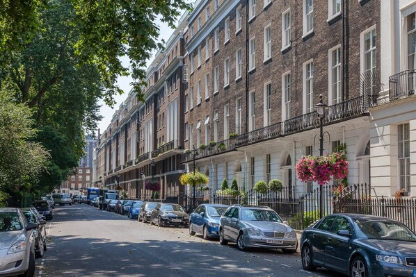 A row of brick townhouses with multiple stories, parked cars along the street, and hanging flower baskets on the balconies.