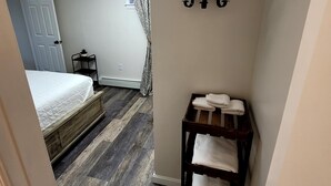Quarto de hotel com cama, um banco de madeira com toalhas, um porta-roupas fixado na parede e uma janela com cortinas.