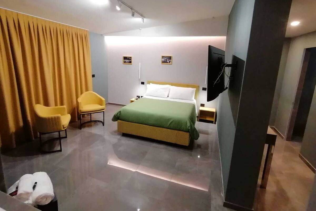 A modern hotel room with a bed, two chairs, a TV, and a large window.