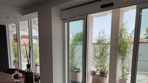 A modern reception area with a stone counter, large sliding glass doors leading to an outdoor space with potted plants and a view of buildings. - X Business (Saviano)