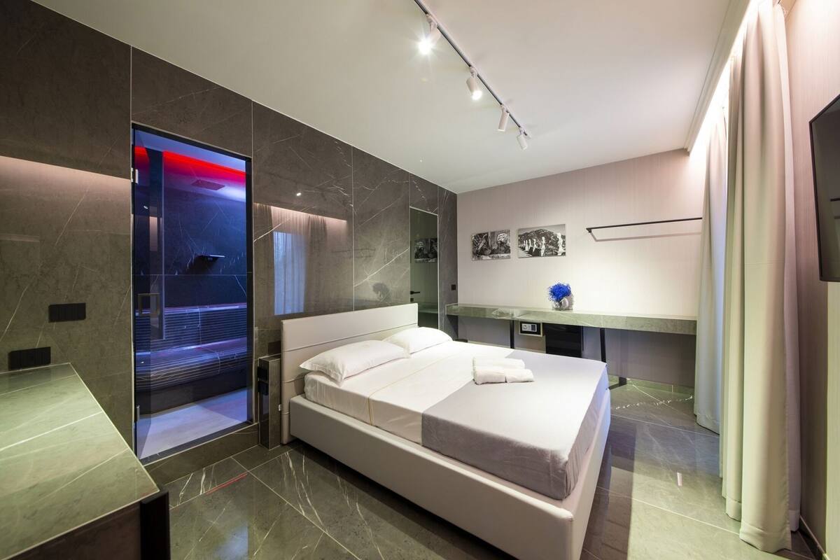 A modern hotel room with a large bed, a vanity with a mirror, and a view of the cityscape through a glass wall.